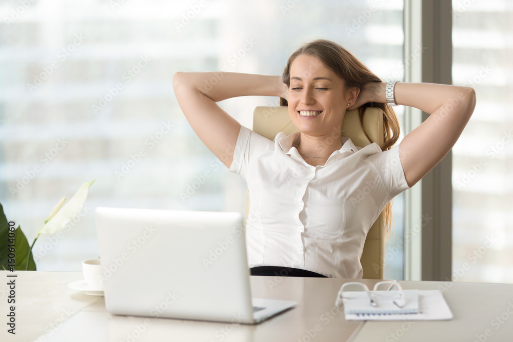 Smiling businesswoman sitting at the desk with hands behind head and looking at laptop screen. Cheerful girl entertains at work with computer. Business lady satisfied watching completed work Smiling businesswoman sitting at the desk with hands behind head and looking at laptop screen. Cheerful girl entertains at work with computer. Business lady satisfied watching completed work