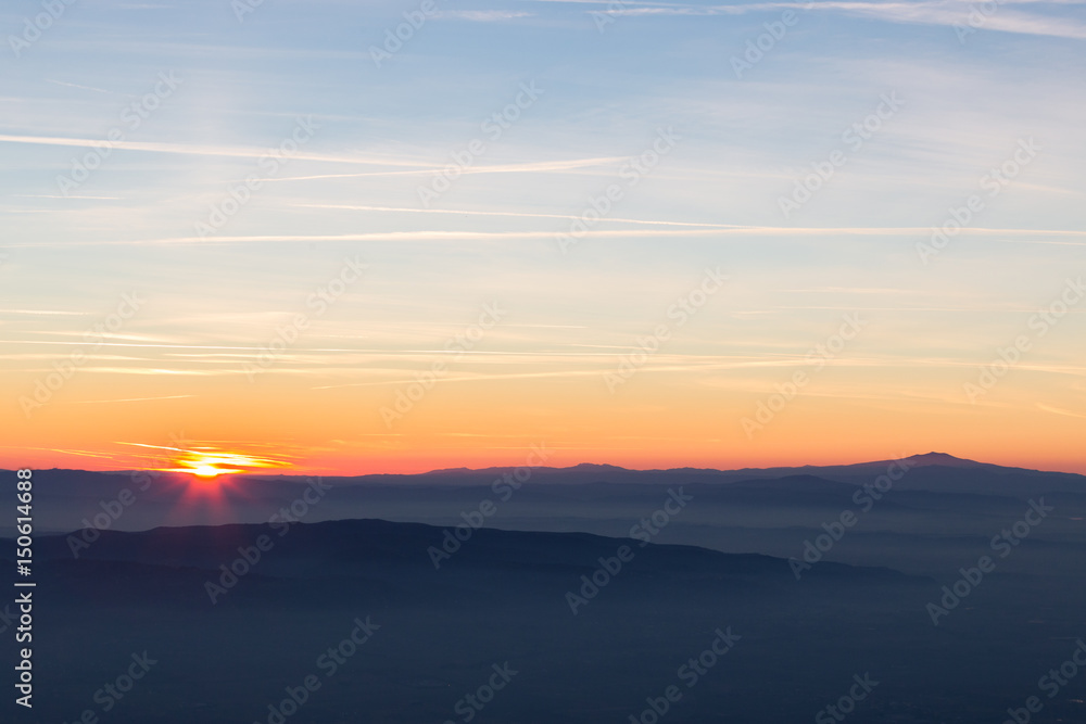 Sun coming down behind some misty mountains, the sky is orange and blue, with many long and thin clouds and jet trails
