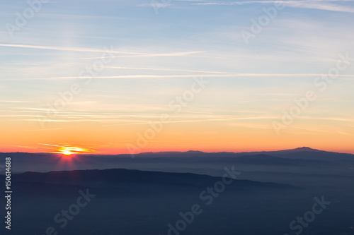 Sun coming down behind some misty mountains, the sky is orange and blue, with many long and thin clouds and jet trails © Massimo