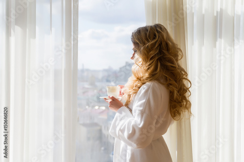 Beauty girl drinking coffee. Beautiful woman opening curtains, looking out the window and enjoying her morning coffee