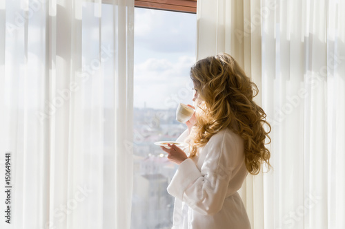 Beauty girl drinking coffee. Beautiful woman opening curtains, looking out the window and enjoying her morning coffee