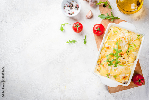Casserole. French cuisine. Homemade potato gratin in a ceramic frying pan for baking. On a white marble stone background. With leaves of fresh arugula, tomatoes, olive oil.  copy space