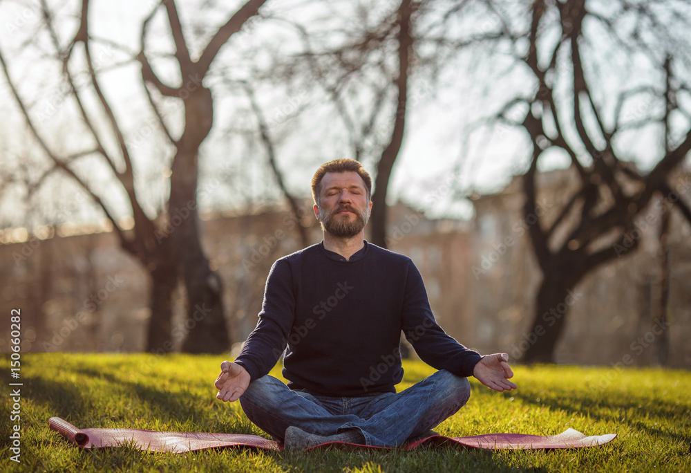 Mature man sitting on yoga mat in comfortable asana relaxing in park. Meditation in city life conditions Mature man sitting on yoga mat in comfortable asana relaxing in park. Meditation in city life conditions