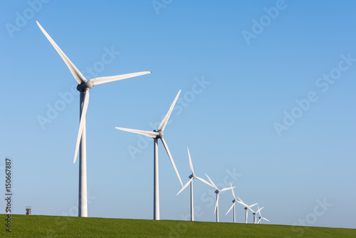 Wind turbines standing along a grassy dyke under a blue sky.