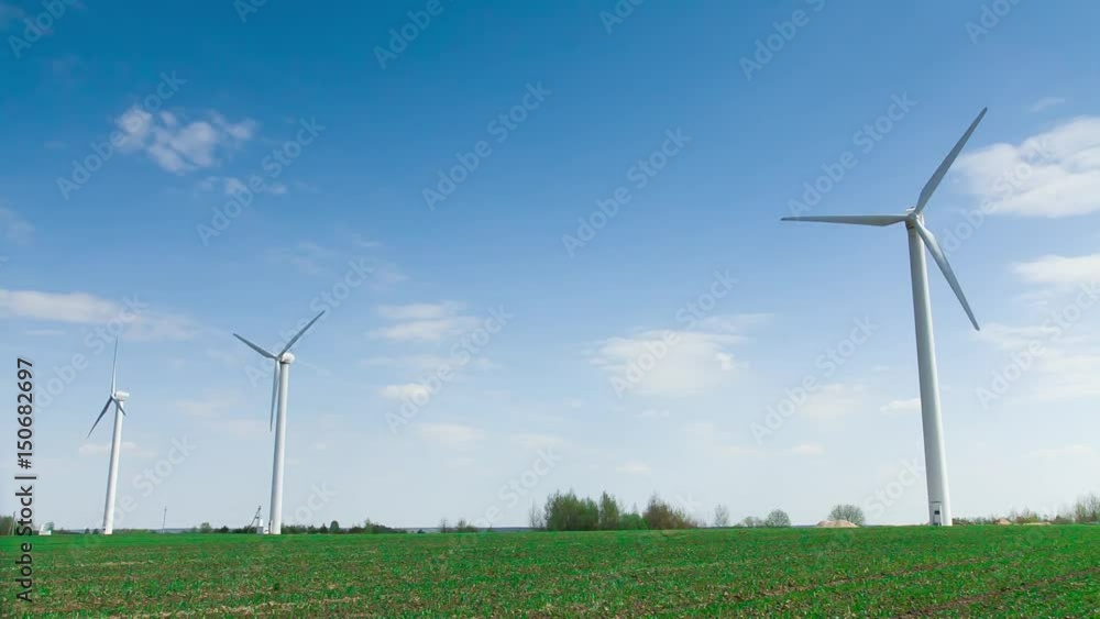 The blades of the three wind stations are spinning against the beautiful blue sky with feathery clouds on a sunny day. Three wind farms is white. Wind energy. Open green field. The blades of the three wind stations are spinning against the beautiful blue sky with feathery clouds on a sunny day. Three wind farms is white. Wind energy. Open green field.
