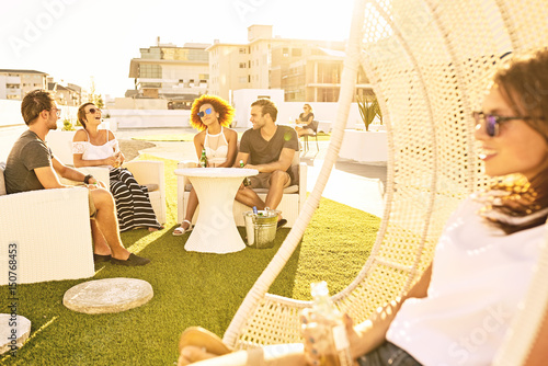 Happy group of attractive friends drinking and socialising outdoors on a green lawn during a warm summer evening during sunset.