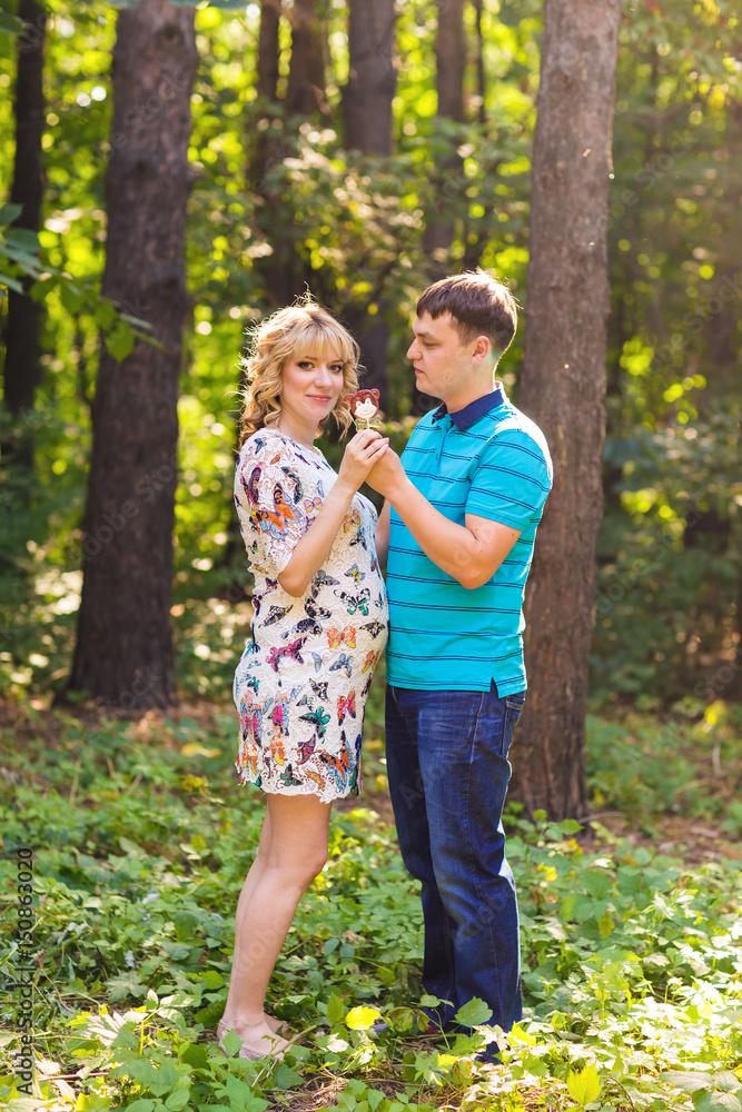Fototapeta premium Pregnancy, family, happiness and fun concept - Man and pregnant woman have fun with candy in the park