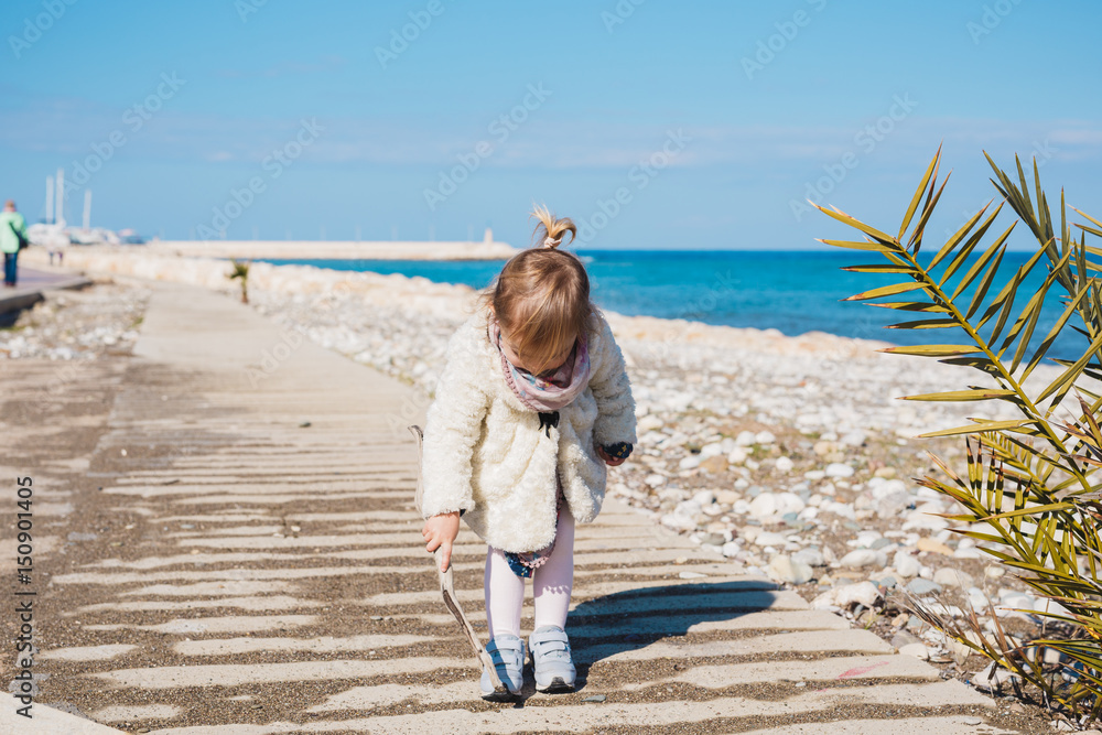 Child, childhood and people concept - Baby girl sits on square road and play with stick near sea beach.