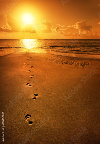 Single footsteps on beach walking towards sun of coastal horizon.