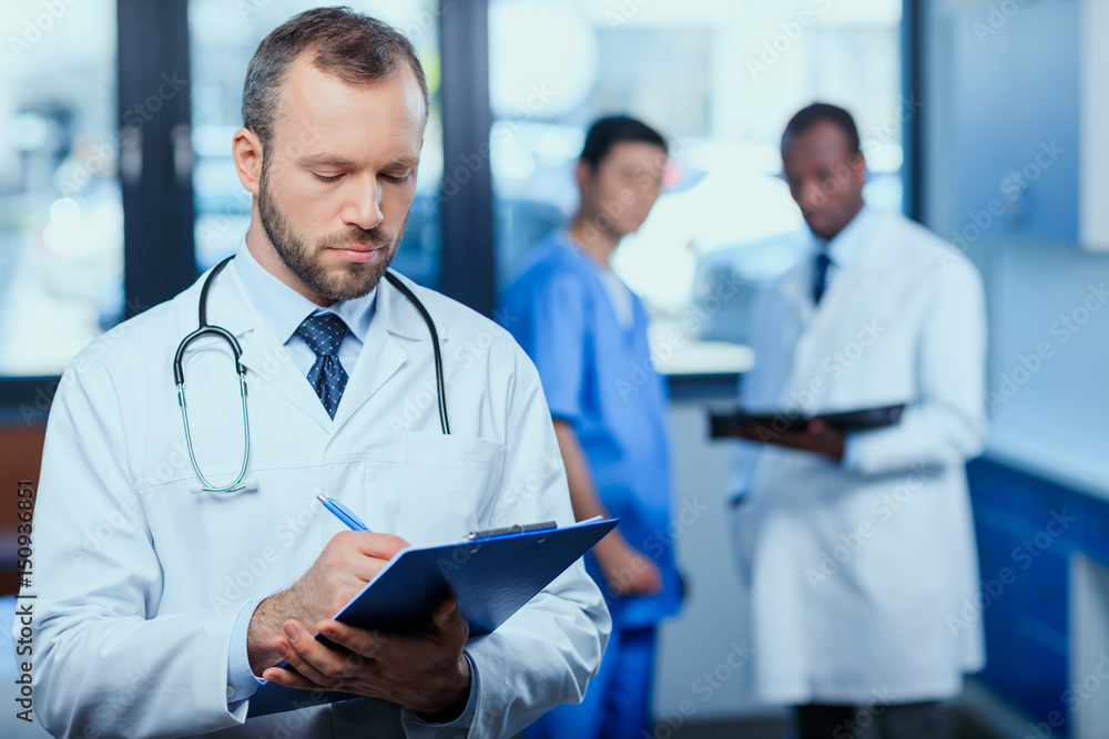 portrait of confident doctor writing in folder in clinic with colleagues behind portrait of confident doctor writing in folder in clinic with colleagues behind