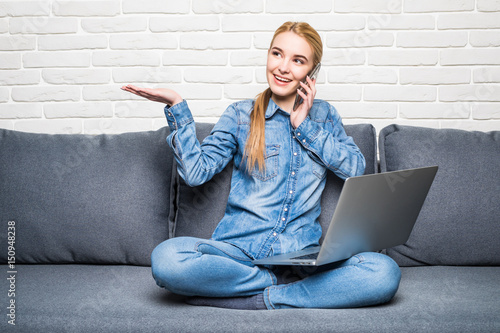 Smiling woman holding a smartphone and calling while using laptop on sofa photo