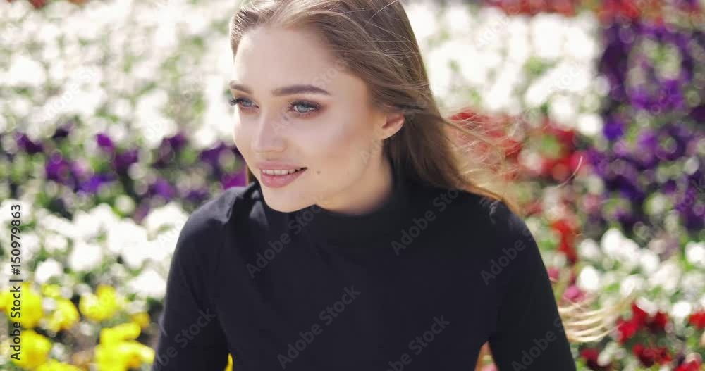 Gentle young woman in casual clothing with hair waving posing on blurred background of vivid flowers Gentle young woman in casual clothing with hair waving posing on blurred background of vivid flowers