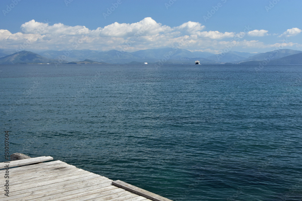 Obraz premium Old wooden dock, distant ferryboat in calm blue sea, mainland on the background, blue sky with white clouds