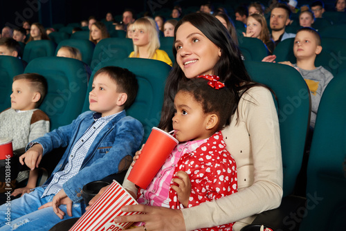 Attractive young woman hugging her cute little daughter smiling joyfully watching cartoon together at the cinema copyspace family parents mother kids childhood positivity leisure concept.