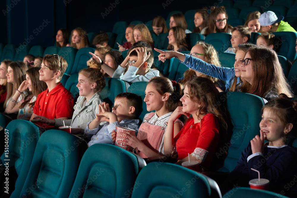 Fototapeta premium Young girls enjoying watching a movie together at the cinema laughing pointing at the screen with their fingers copyspace carefree positive emotions expressing vitality lifestyle leisure entertaining.