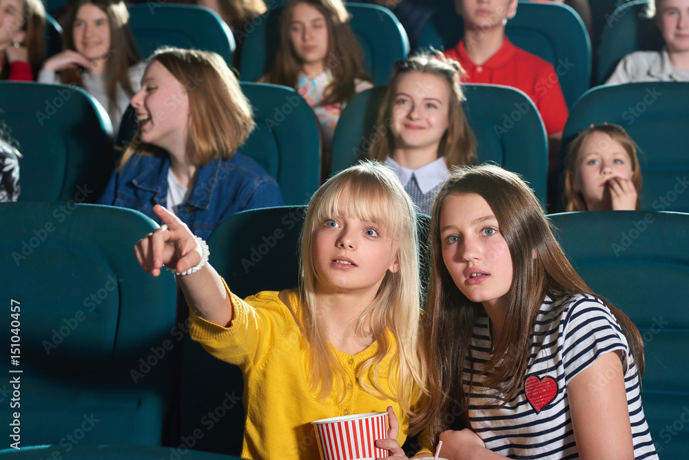 Young pretty girl looking surprised pointing at the movie screen at the cinema showing her friend something copyspace entertainment expressive emotions shock surprised hobby premiere tickets concept.