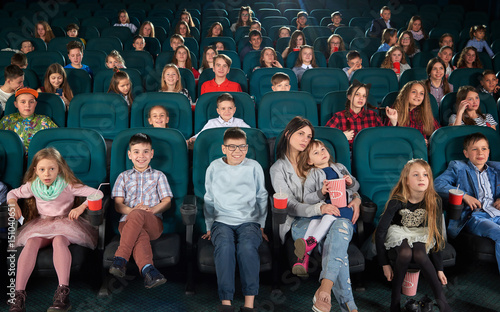 Full length shot of a woman enjoying watching a movie at the cinema with her little daughter in an auditorium full off children youth lifestyle leisure entertainment people activity concept.