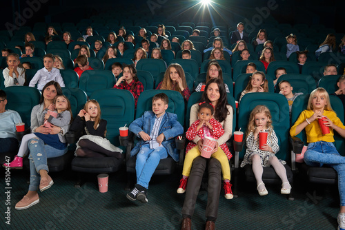 Full length shot of a cinema auditorium full of kids enjoying cartoon premiere. Young woman with her daughter on her lap watching a movie together family children entertainment weekend concept.