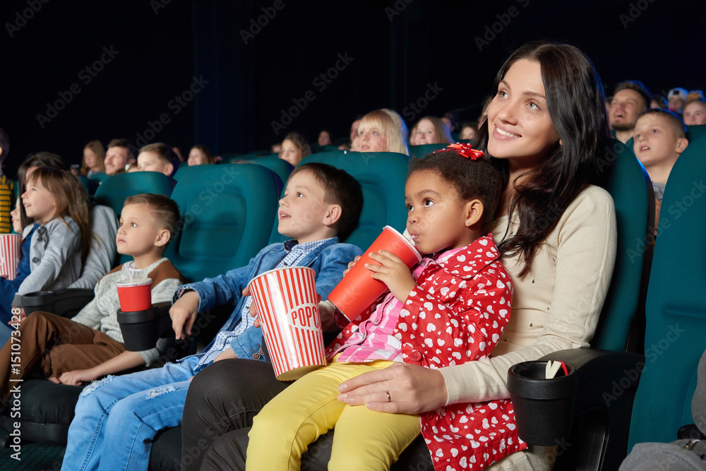 Fototapeta premium Young beautiful woman smiling holding her young daughter on the lap enjoying movie premiere at the cinema copyspace happiness living people leisure activity weekend togetherness concept.