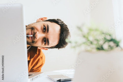 Funny close up shot of happy and smiling young male executive hidden behind laptop at office with pencil on mouth. Young European businessman working at home. Handsome salesman have secret of succes.  photo