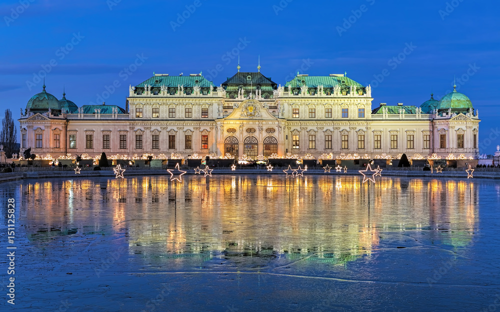 Upper Belvedere Palace with Christmas Village reflecting in the pond covered with wet ice in evening, Vienna, Austria