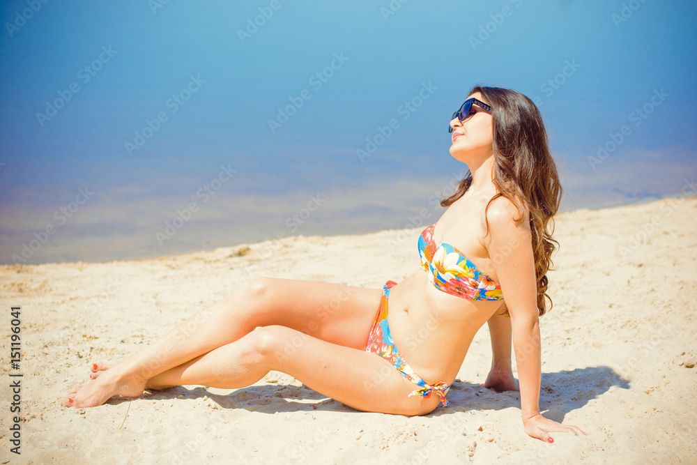 Beautiful spanish brunette with long hair on the beach in a motley swimsuit on a warm sand near the blue water. A woman rests and relaxes with a view of the open air Beautiful spanish brunette with long hair on the beach in a motley swimsuit on a warm sand near the blue water. A woman rests and relaxes with a view of the open air