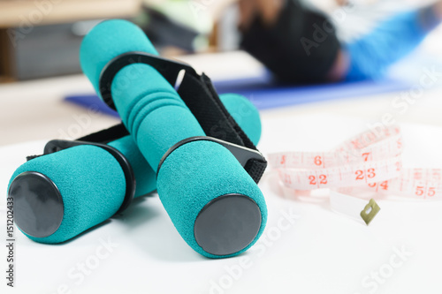 Fitness at home, a pair of green dumbbells and messuring tape in the foreground and man doing sit-ups on the background. photo