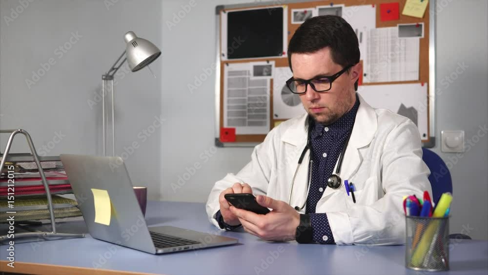 A young doctor in a white lab coat and a stethoscope on the neck examines X-rays on a mobile phone in the private clinic's office. A man heals patients using a laptop and other gadgets A young doctor in a white lab coat and a stethoscope on the neck examines X-rays on a mobile phone in the private clinic's office. A man heals patients using a laptop and other gadgets