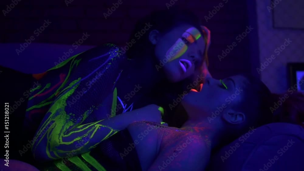Two beautiful women lying on sofa very close to each other in fluorescent clothing under UV black light. One woman inhaling hookah Two beautiful women lying on sofa very close to each other in fluorescent clothing under UV black light. One woman inhaling hookah