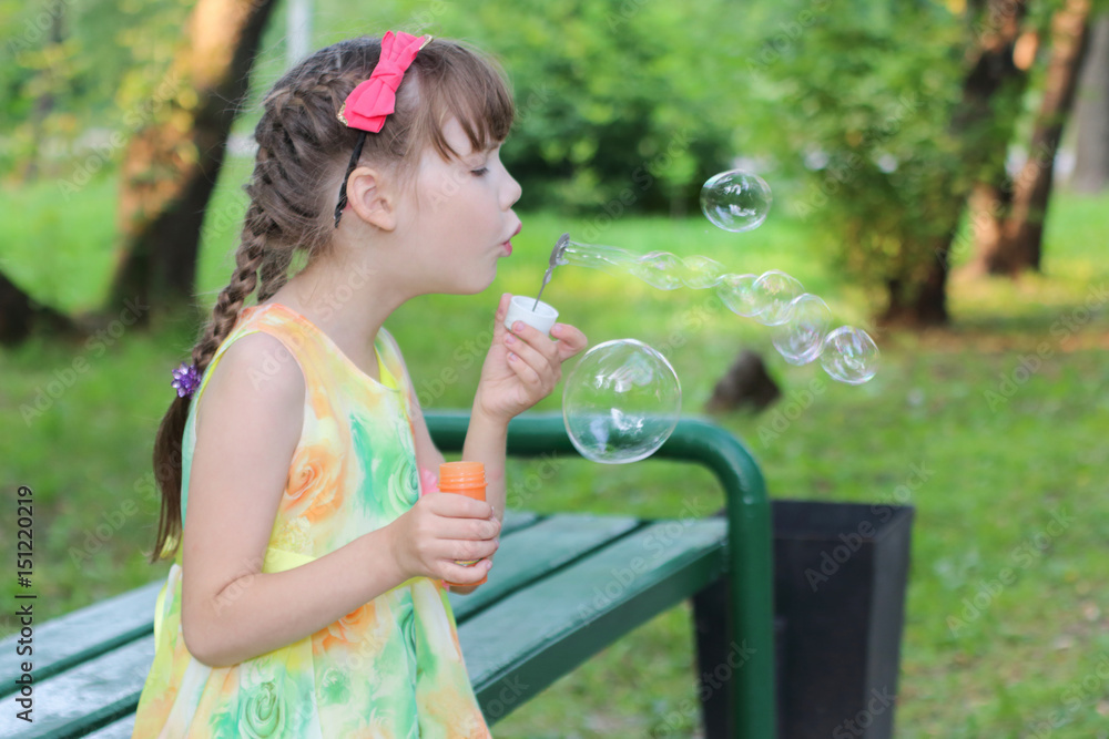 Little girl with pigtails and a cute bow on the head with blow bubbles sitting on the bench Little girl with pigtails and a cute bow on the head with blow bubbles sitting on the bench