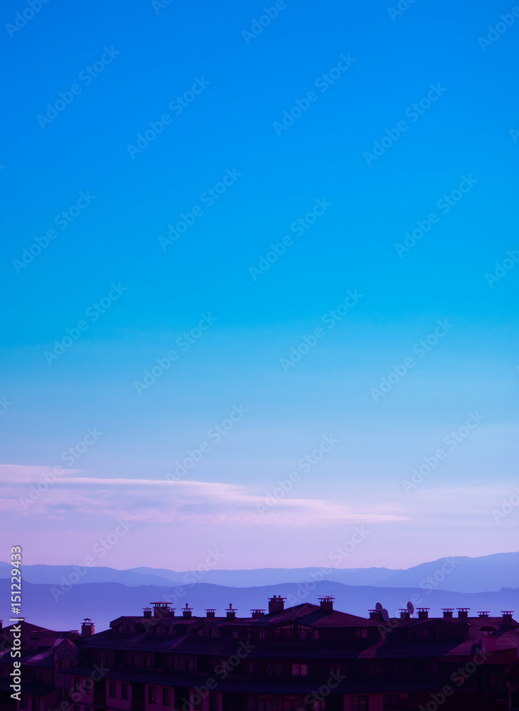 Fototapeta premium Photo depicting a beautiful European mountain landscape with city chalet roofs silhouettes in front. European alpine mountains with snow peaks on a clean blue sky background.