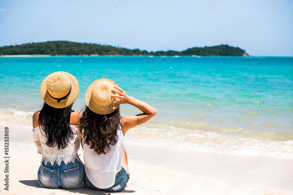 Two beautiful young hipster girls sit on the seashore, the ocean, look at the turquoise water, enjoy the sun, are dressed in white T-shirts, denim shorts, vacation in a tropical country, travel
