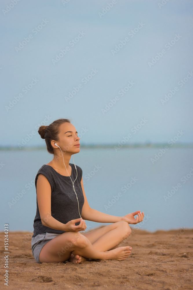 Young beautiful woman meditating on the beach in the morning at the dawn of the sun in a gray T-shirt and shorts