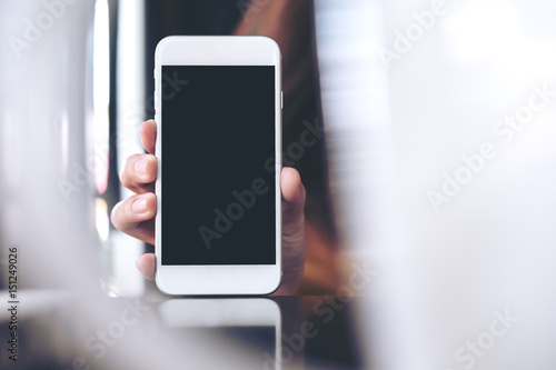 Mockup image of hand holding white mobile phone with blank black screen in cafe and blur foreground