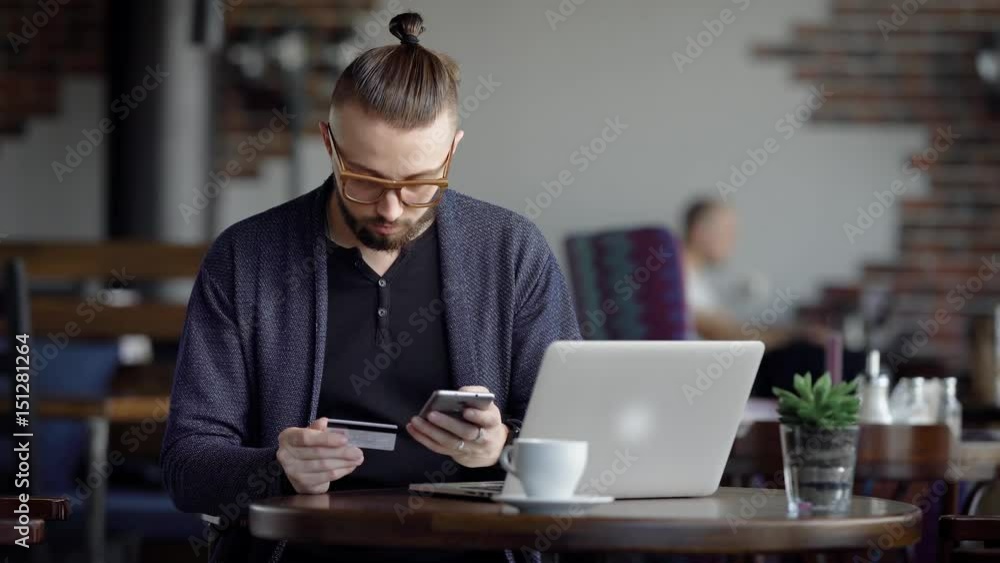 A young man sits in a cafe and makes an online purchase of stylish clothes in an online store, a person uses a credit card and a mobile phone for payment, on the desk is a laptop A young man sits in a cafe and makes an online purchase of stylish clothes in an online store, a person uses a credit card and a mobile phone for payment, on the desk is a laptop