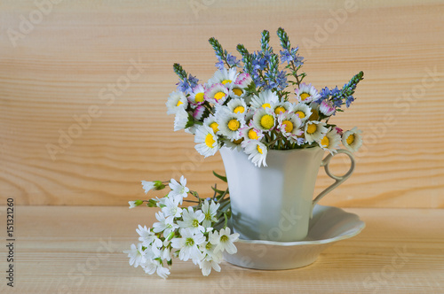 Bouquet of white of field flowers in a cup with a plate on a wooden background