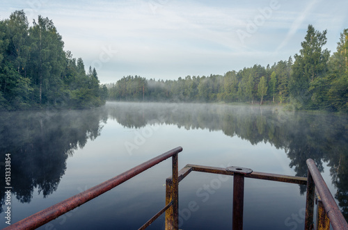 Early morning on the forest lake. Metal bridge in foreground. Fog. Reflection on water.