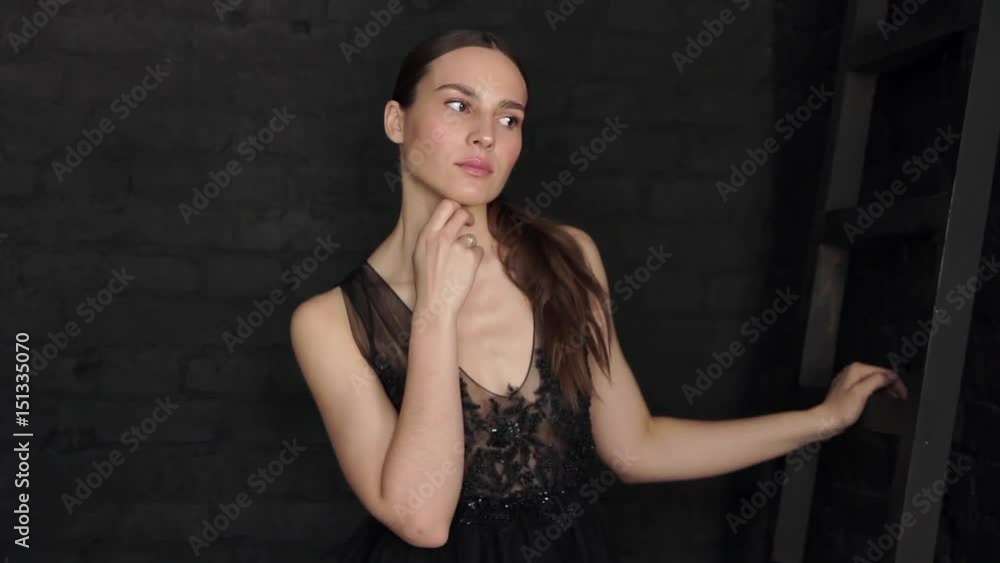 Beautiful brunette in a black dress posing and looking at the camera on a black wall background. Beautiful brunette in a black dress posing and looking at the camera on a black wall background.