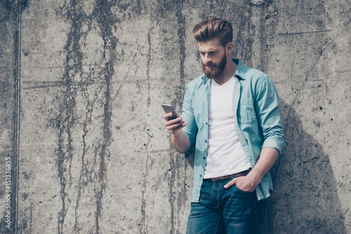 Pensive brutal young red bearded guy is typing a message outdoors. He is very stylish, wearing jeans casual clothes