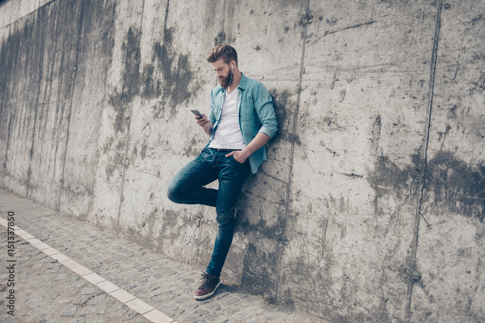 Hot young stylish guy is standing near the concrete wall outside and browsing on his pda. He has a fashionable hairstyle and beard, wearing casual clothes Hot young stylish guy is standing near the concrete wall outside and browsing on his pda. He has a fashionable hairstyle and beard, wearing casual clothes