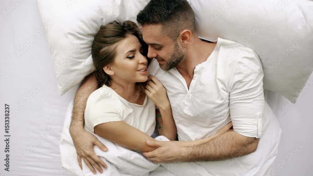 Young beautiful and loving couple kiss and hug into bed while waking up in the morning. Top view of attractive man kissing and talking his smiling wife Young beautiful and loving couple kiss and hug into bed while waking up in the morning. Top view of attractive man kissing and talking his smiling wife