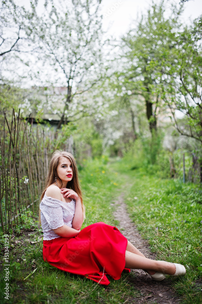 Portrait of sitting beautiful girl with red lips at spring blossom garden on green grass, wear on red dress and white blouse. Portrait of sitting beautiful girl with red lips at spring blossom garden on green grass, wear on red dress and white blouse.