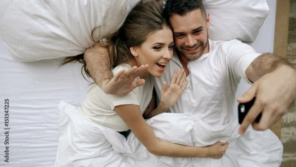 Young beautiful and loving couple take selfie picture on smartphone camera and kiss while lying in bed at the morning Young beautiful and loving couple take selfie picture on smartphone camera and kiss while lying in bed at the morning