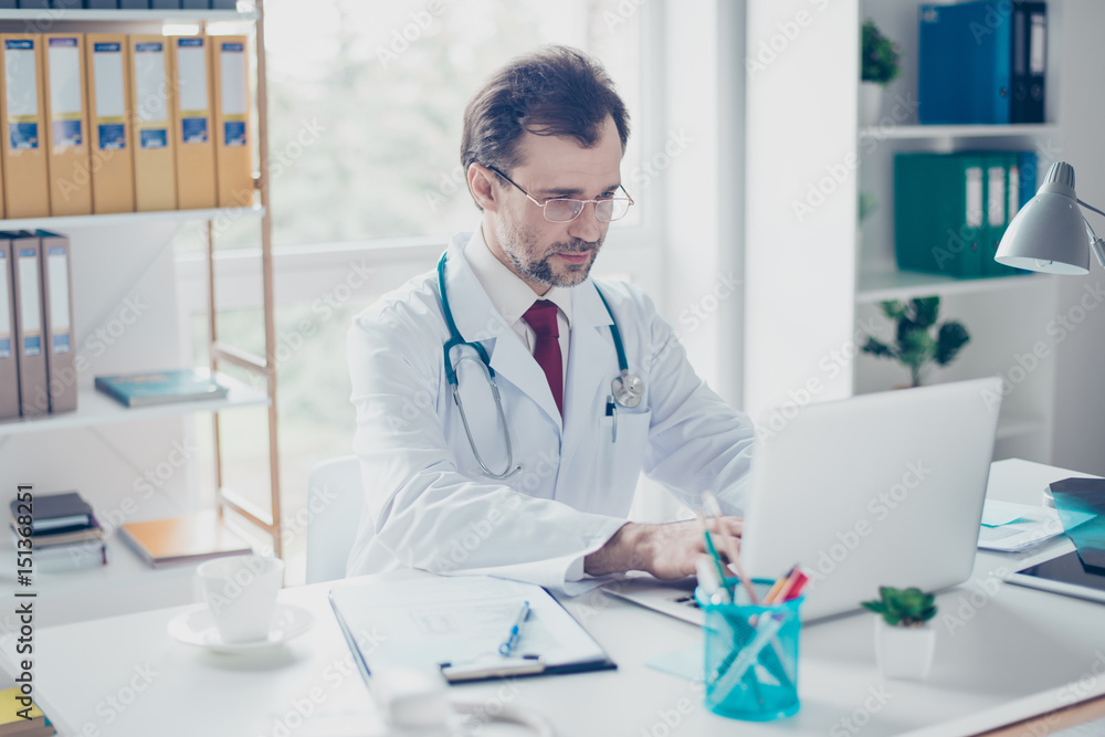 Serious doctor is browsing info on hia computer. He is in a white coat, glasses, sitting in modern office Serious doctor is browsing info on hia computer. He is in a white coat, glasses, sitting in modern office