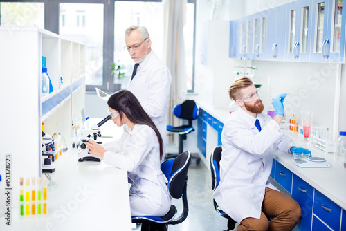 Teambuilding concept. Three workers of laboratory are ckecking the analysis. They look stylish, wearing labcoats. The lab is bright white and blue photo