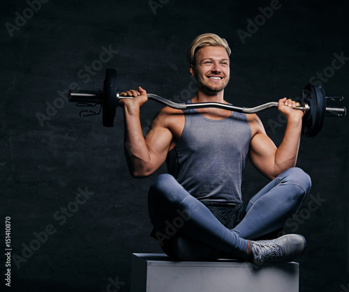 Smiling blond sporty male sits on a white box with crossed legs and holds barbell. © Fxquadro