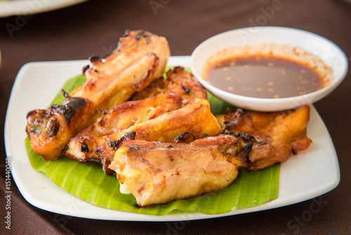 Grilled chicken laid on banana leaves with spicy sauce in the white dish.