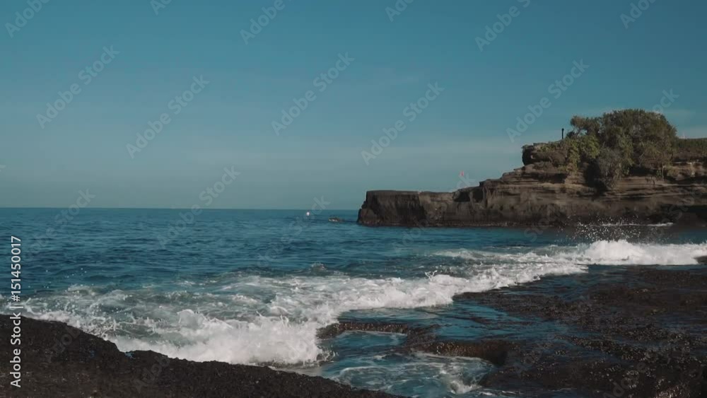 Blue calm water small ocean waves washing and splashing against dark rocks, cliff in background. Shot with Sony a7s on slider on sunny day with blue sky Blue calm water small ocean waves washing and splashing against dark rocks, cliff in background. Shot with Sony a7s on slider on sunny day with blue sky