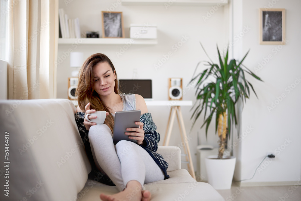Girl drinking morning coffee and using tablet while siting on couch Girl drinking morning coffee and using tablet while siting on couch