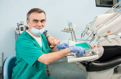 Portrait of senior male dentist with dental equipment in the dental office. Doctor wearing mask  green uniform and blue gloves. On the background blurred patient. Dentistry